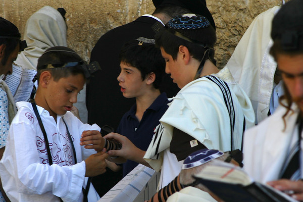 Jonathan Siegel (JSBird) > Youngsters putting on T'fillin. These are boxes with prayers in them, wrapped with leather cords around the arm and the skull. This practice comes from Deuteronomy: in the same passage that commands us to revere the words of God by putting them "on the doorposts of our house" (also known as a Mezuza), the verse continues by commanding us to put them (God's words) before our eyes and upon our arms.